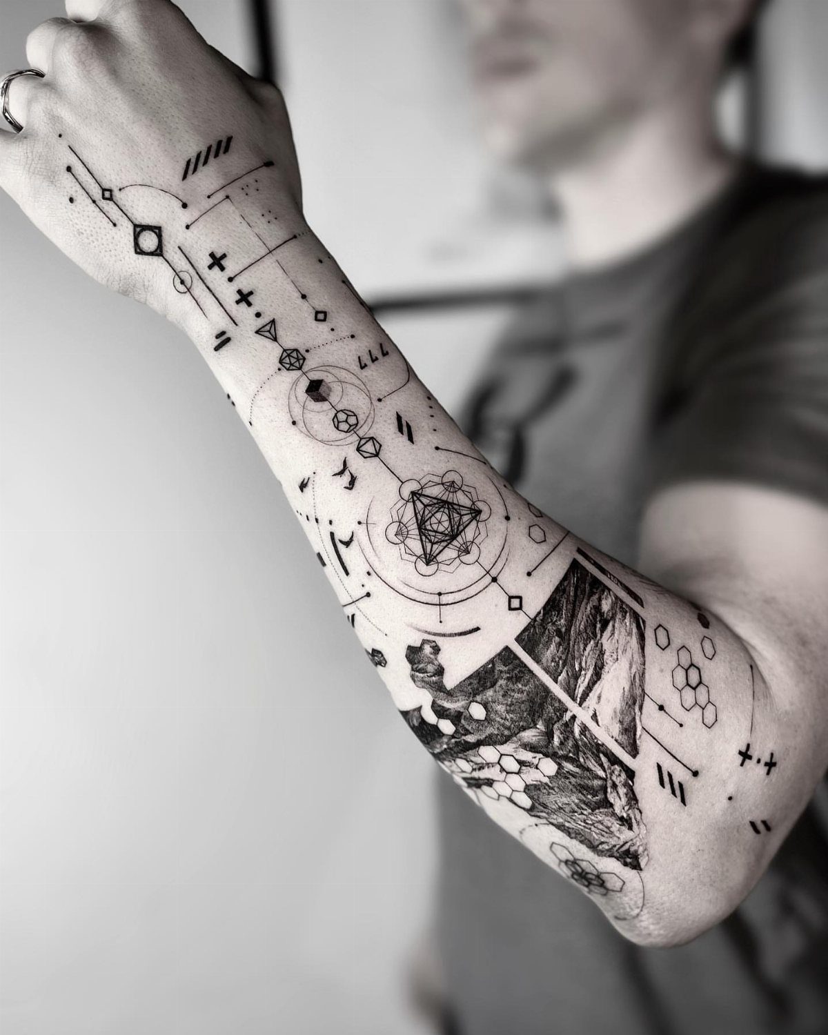 Forearm tattoo with intricate geometric patterns, lines, and a small landscape scene.