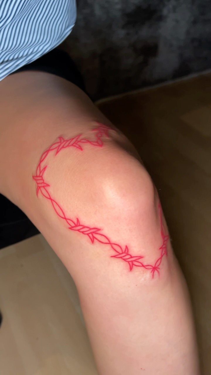Barbed wire heart tattoo in red ink on a person's knee.
