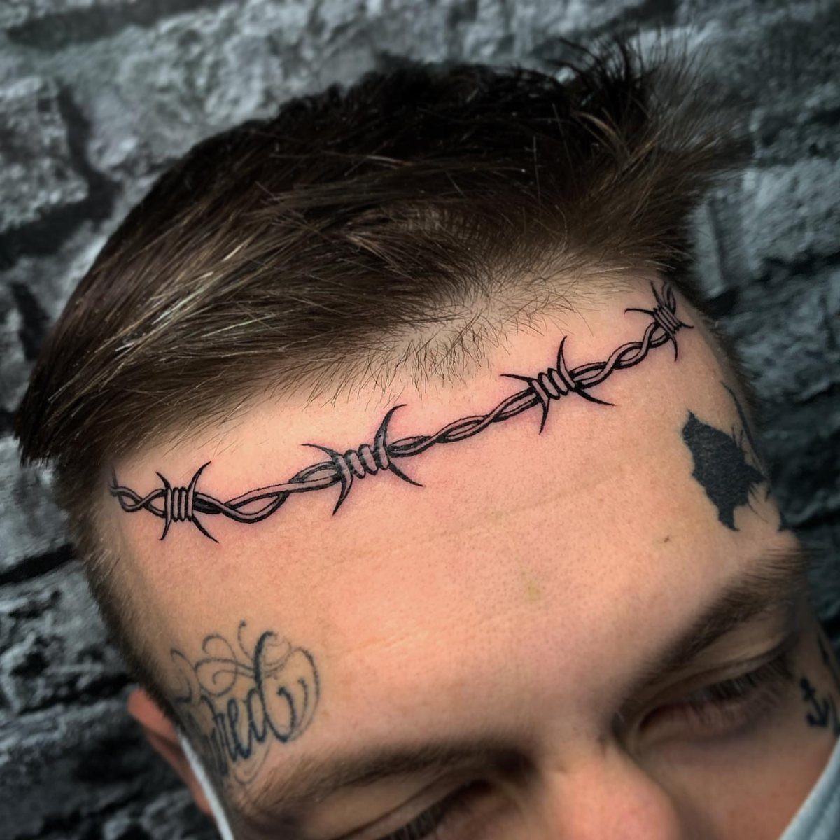Barbed wire tattoo stretching across a man's forehead against a grey stone wall.