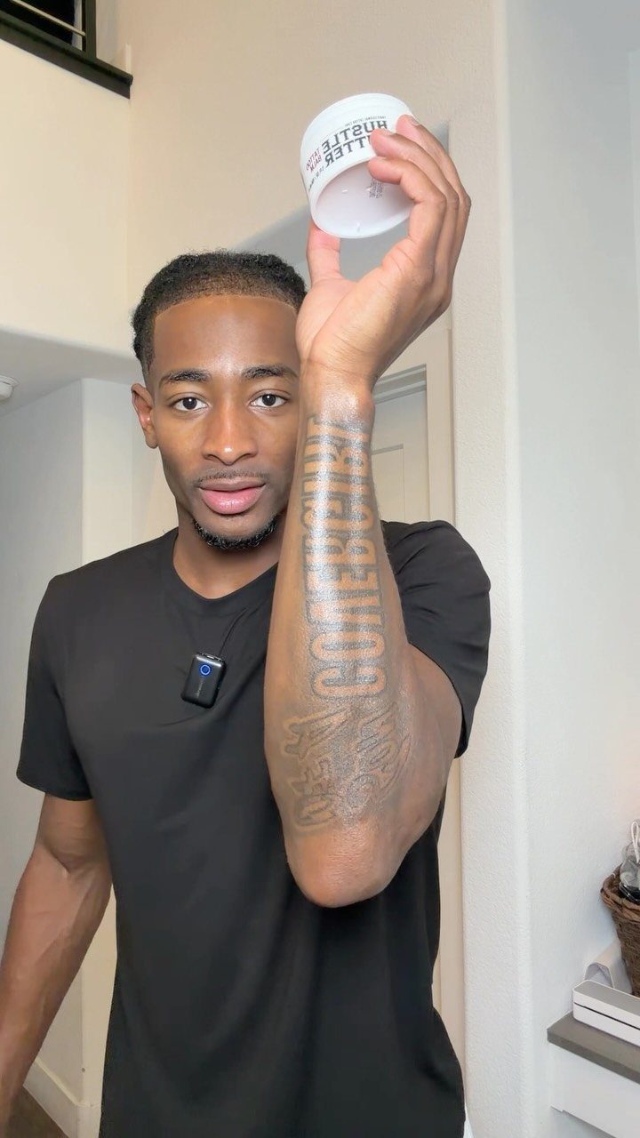 Man with brown skin holding a cream container over his tattooed forearm.