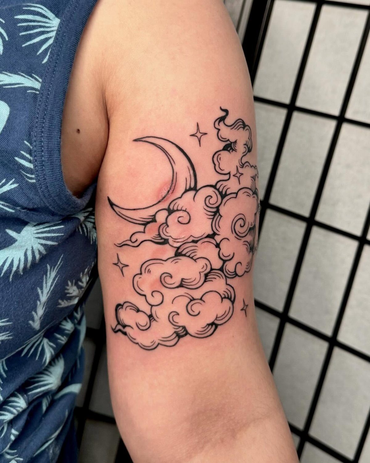 Crescent moon, fluffy clouds, and stars tattoo on a person's upper arm.