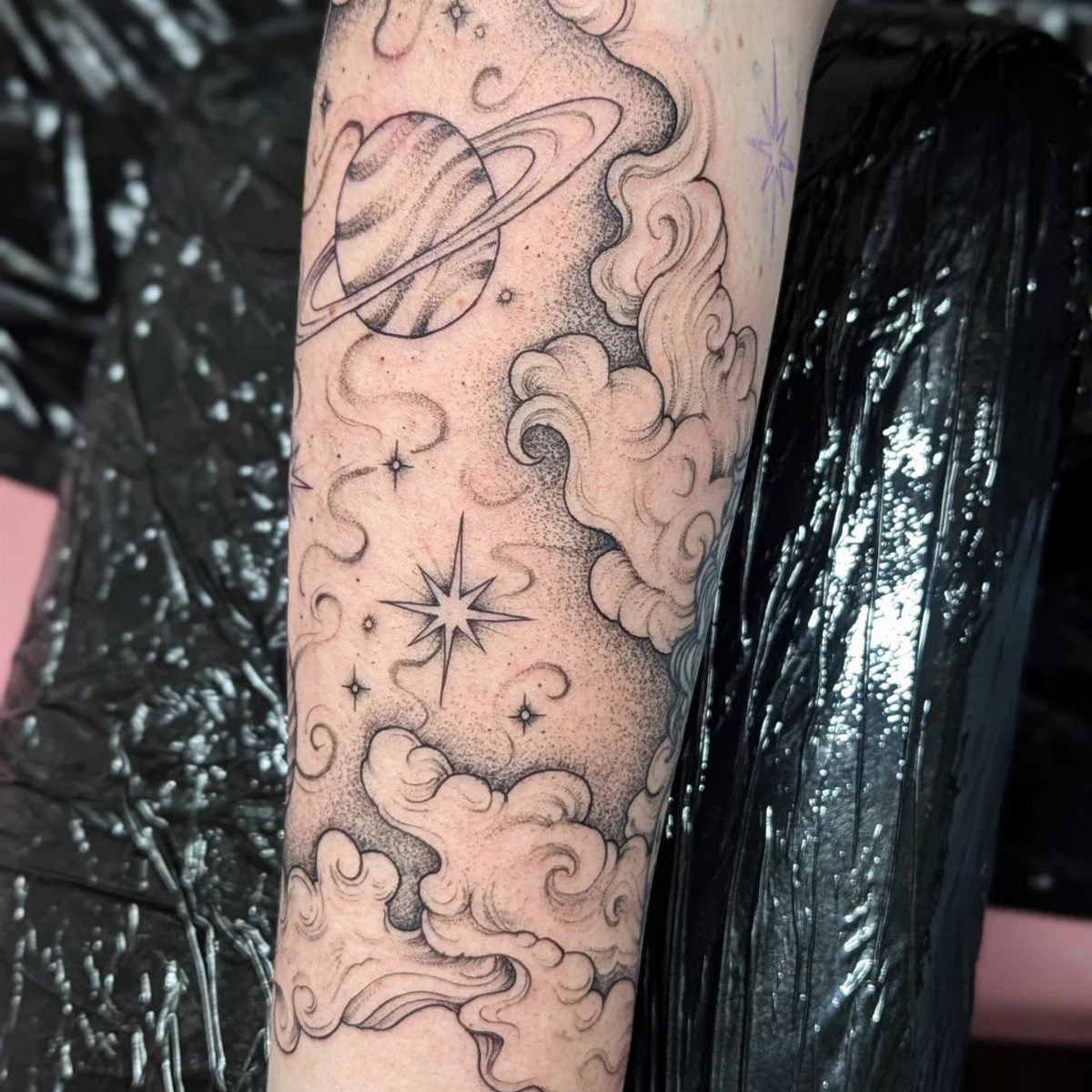 Tattoo of fluffy clouds, stars, and a ringed planet on a person's arm.