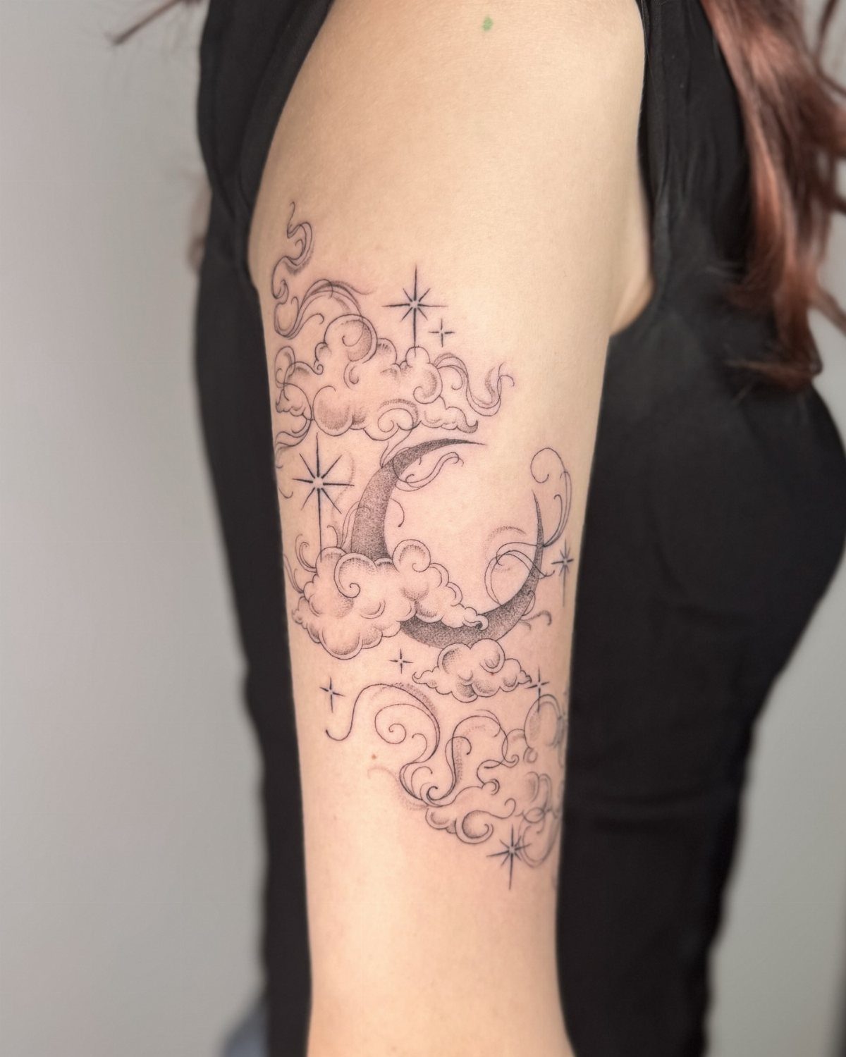 Crescent moon tattoo with clouds and stars on a person's upper arm.