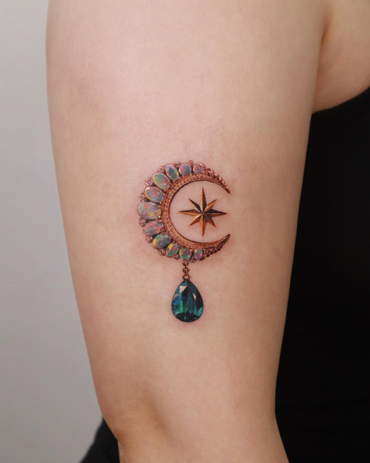 Crescent moon tattoo with iridescent gems and a hanging blue teardrop gemstone.