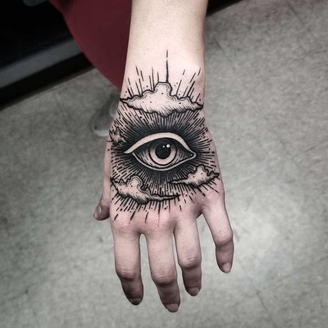 Black ink tattoo of an eye surrounded by dark clouds on a hand.