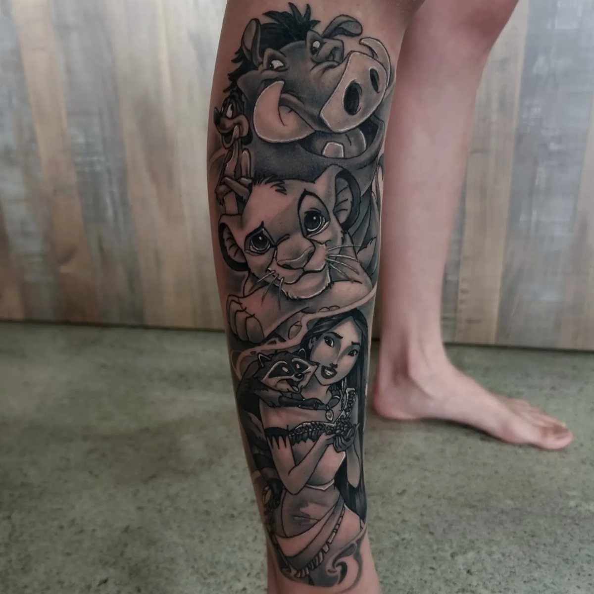 Black and grey tattoo of Pumbaa, Simba, and Jasmine on a lower leg.