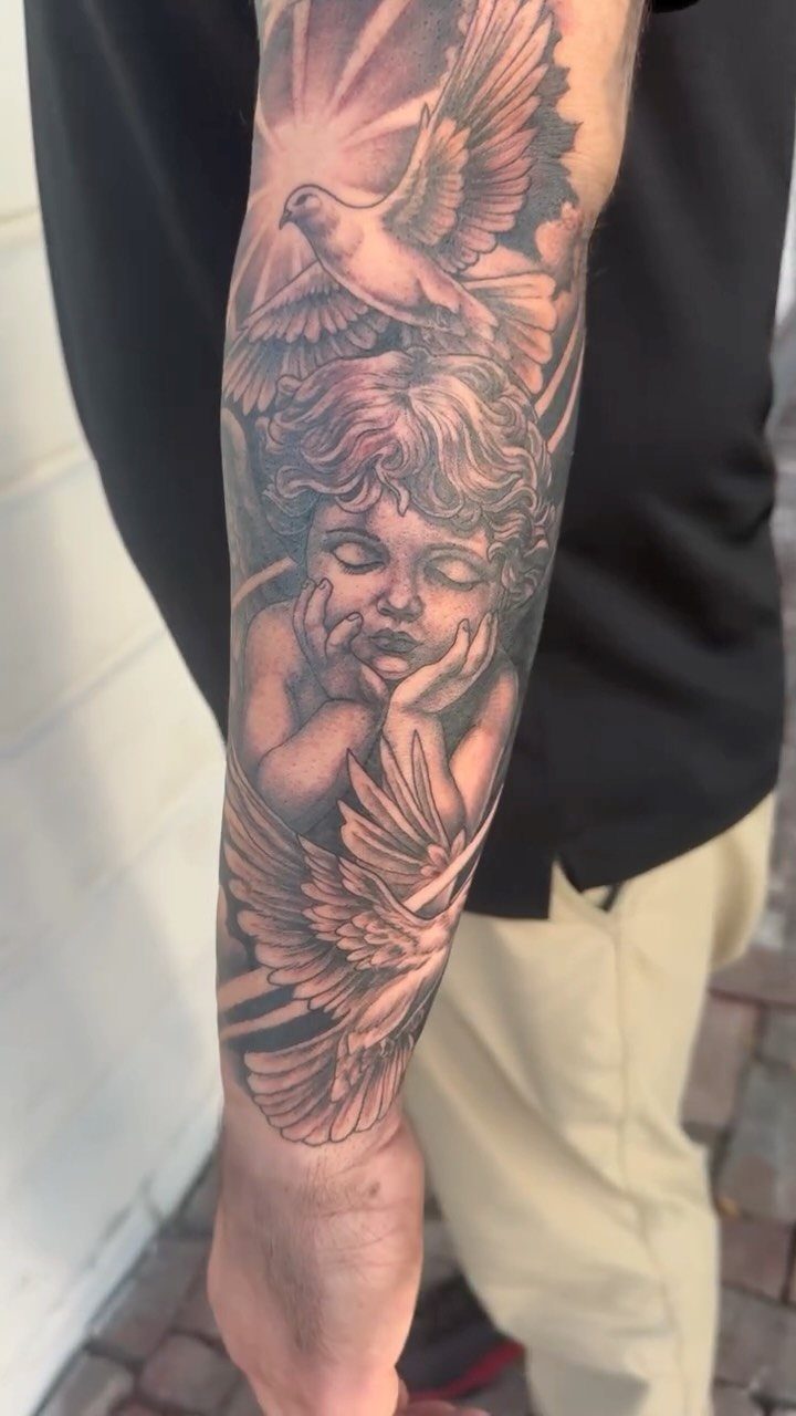 Black and grey forearm tattoo of a sleeping cherub and two doves.