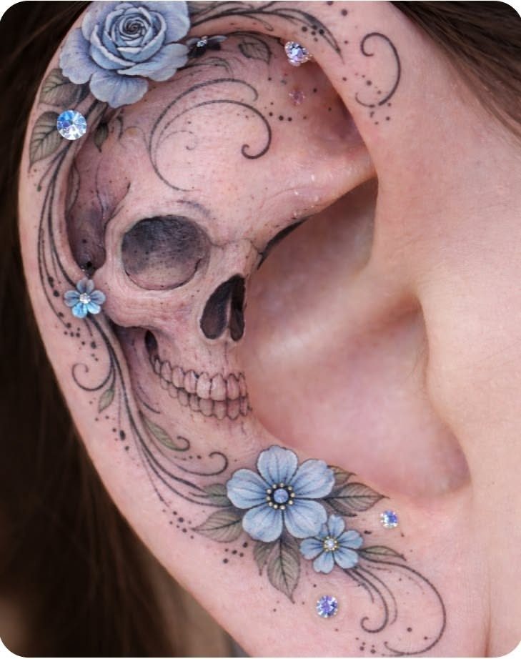 Skull tattoo on an ear surrounded by light blue flowers and swirling lines.