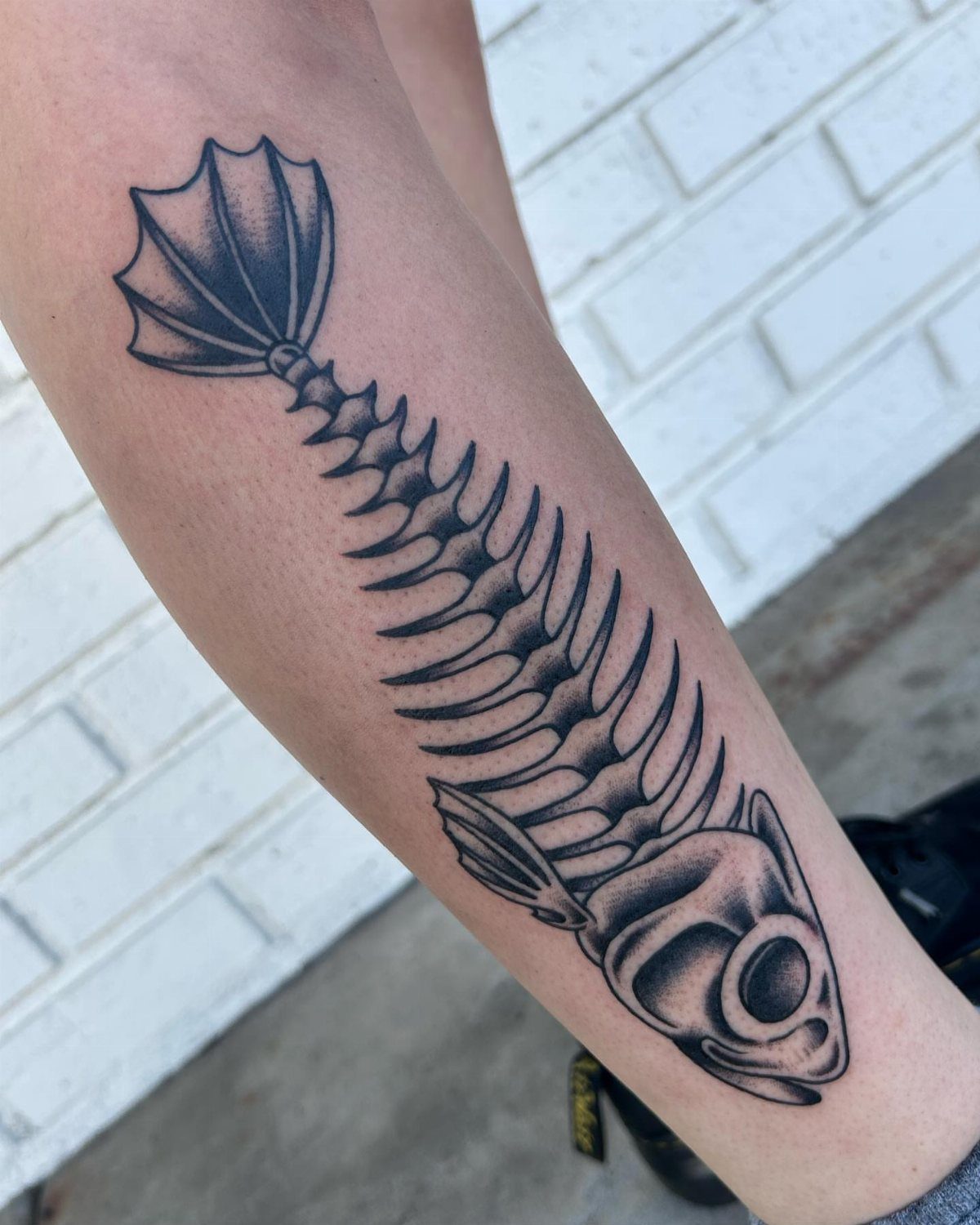 Fish skeleton tattoo on a forearm featuring detailed fine-lined fins and a skull.