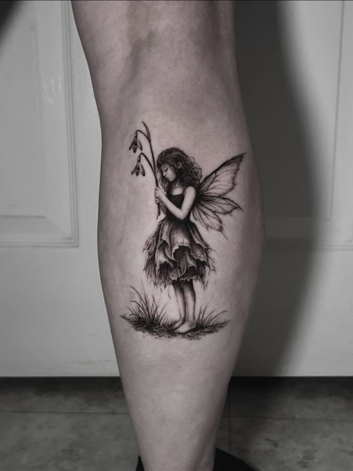 Slender fairy tattoo with ornate wings and a leaf-style ruffled skirt on a leg.