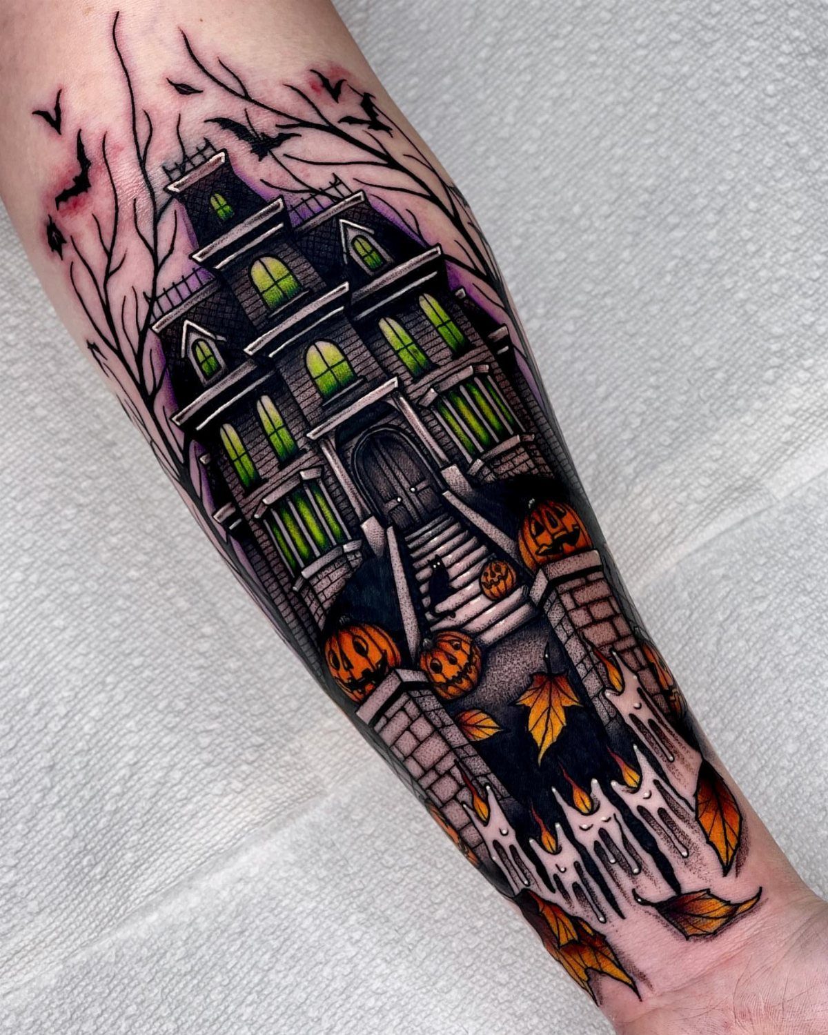 Haunted house tattoo on forearm with glowing green windows and carved pumpkins.