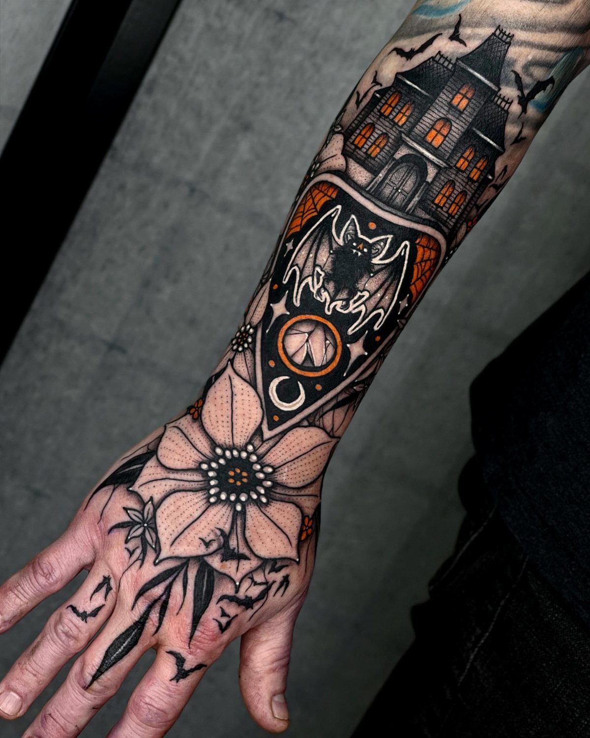 Haunted house tattoo on a forearm with a bat and floral designs.