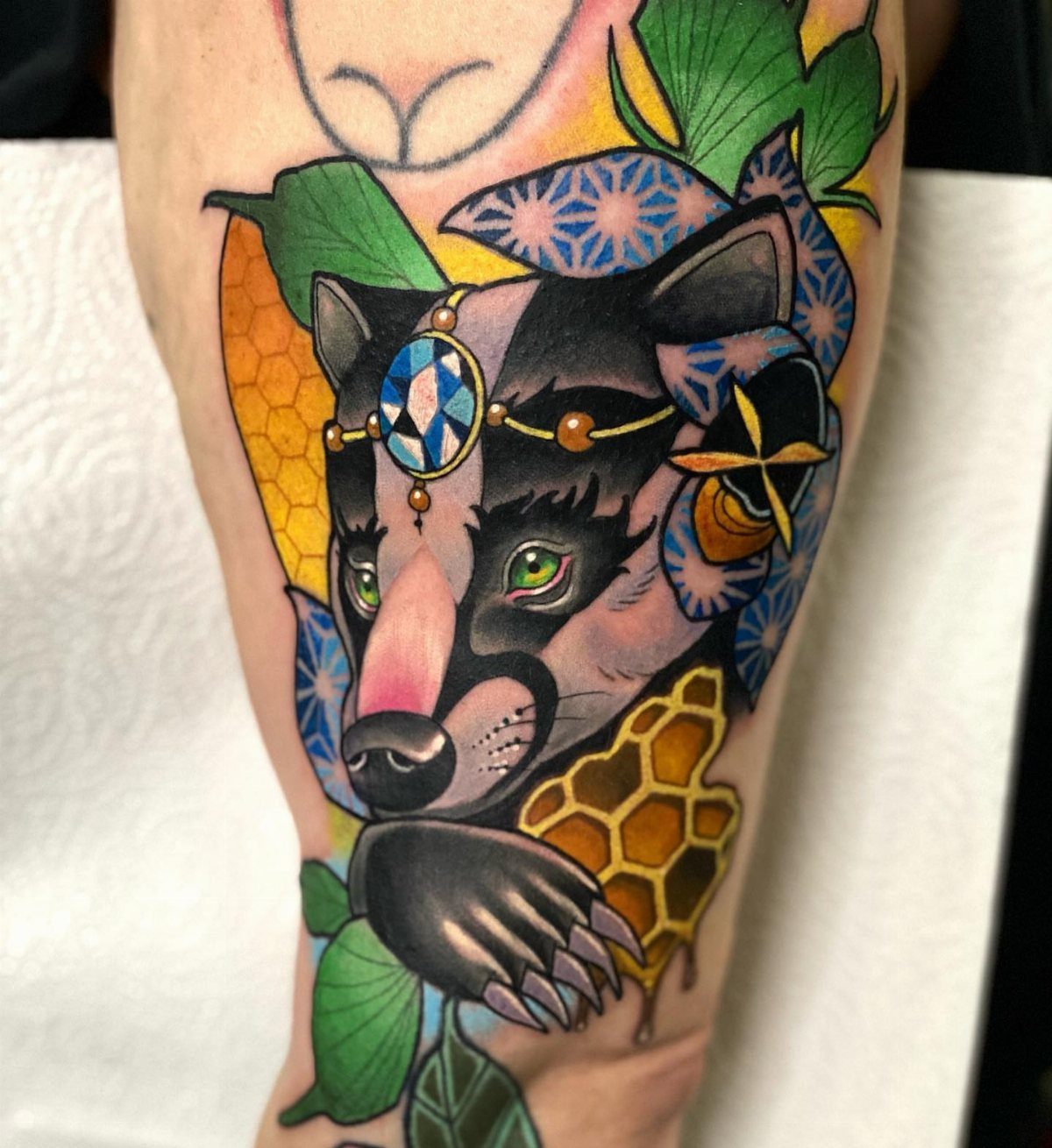 Honey badger tattoo with a jeweled headpiece, honeycomb, and green leaves on skin.