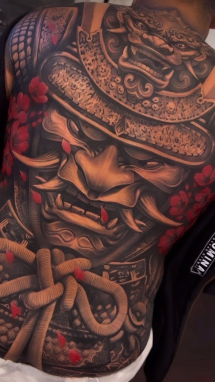 Detailed samurai mask tattoo on a person's back with red floral accents.