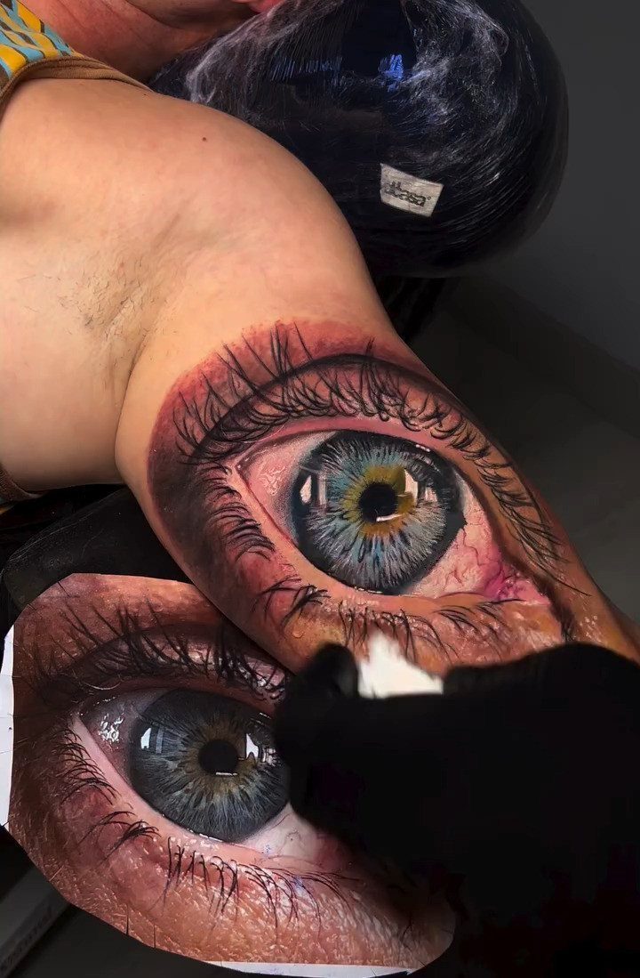 Hyper-realistic eye tattoo on skin next to a reference photograph of an eye.