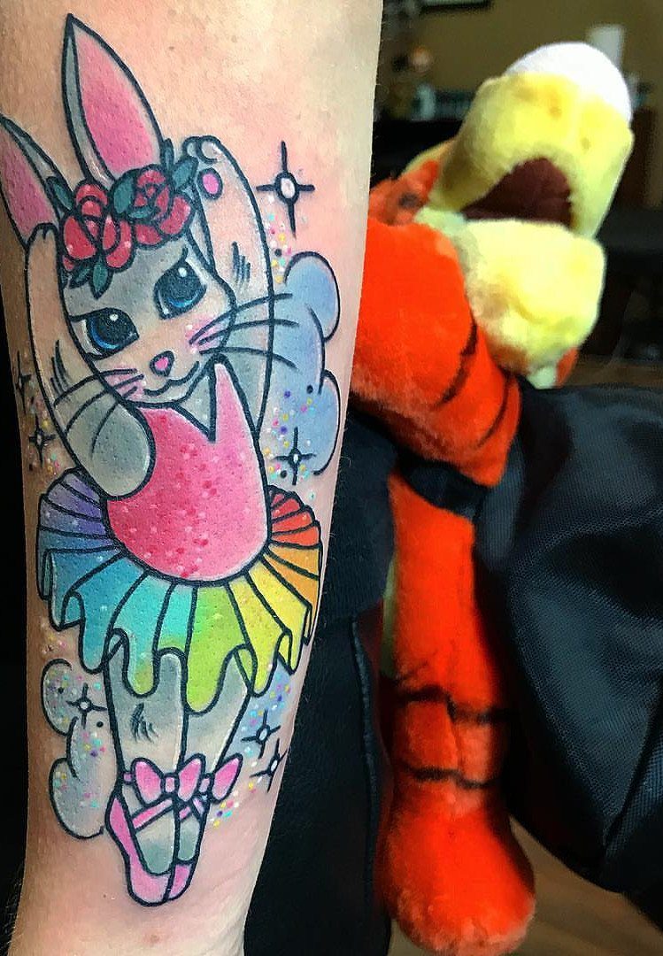 Ballerina bunny tattoo with rainbow tutu and flower crown on a person's arm.