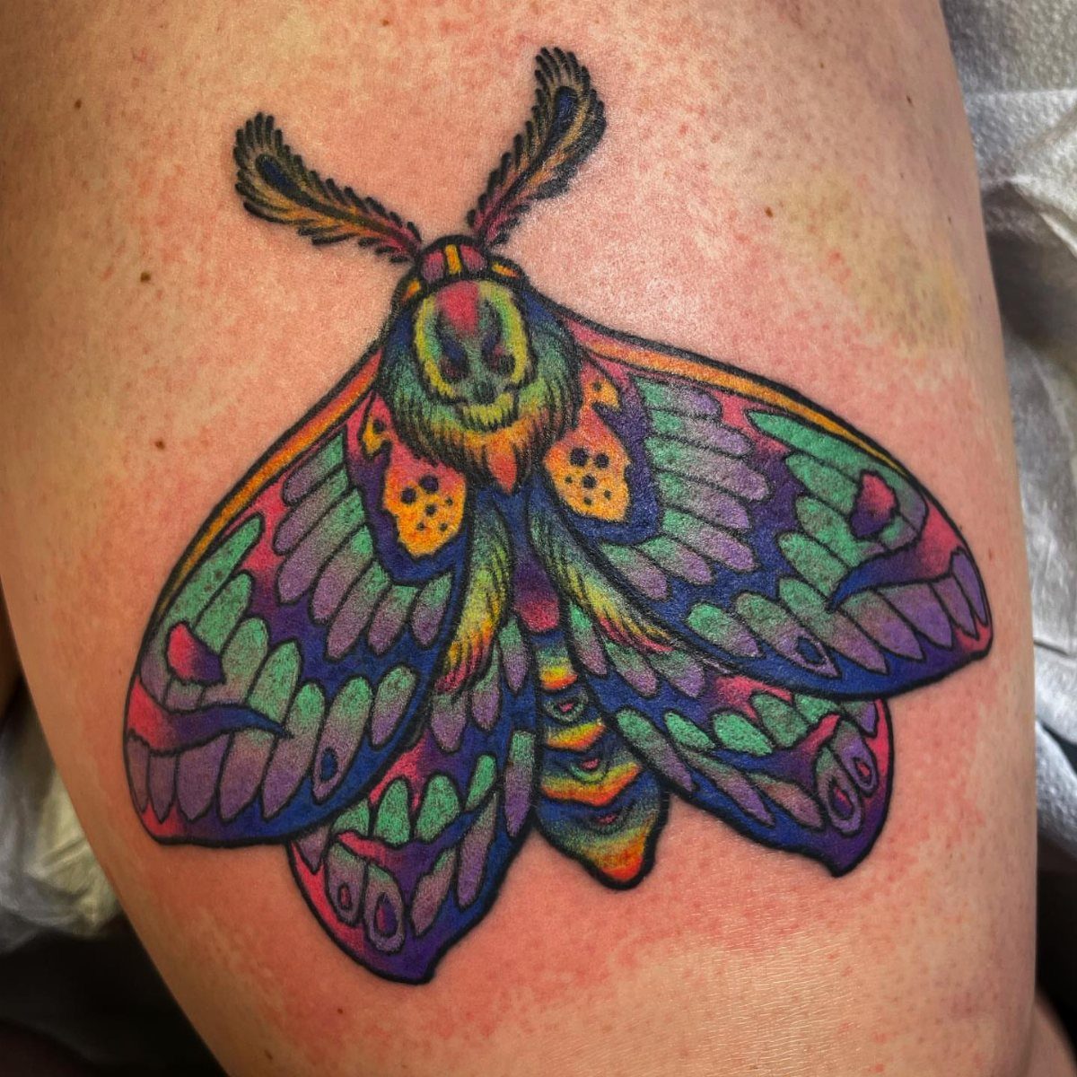 Rainbow colored moth tattoo with vibrant purple, blue, and green wings on skin.