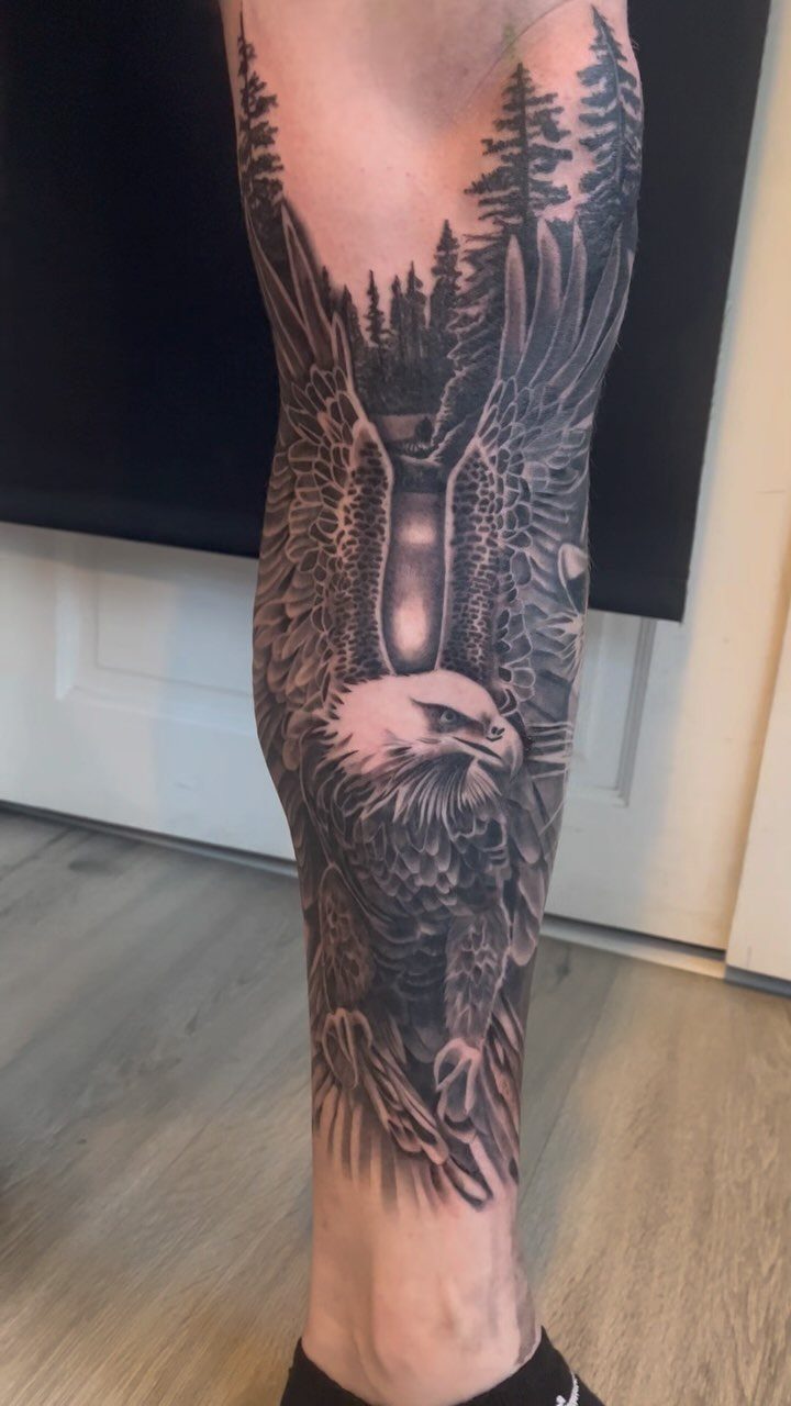 Detailed black and grey eagle tattoo on a man's leg with forest scenery.