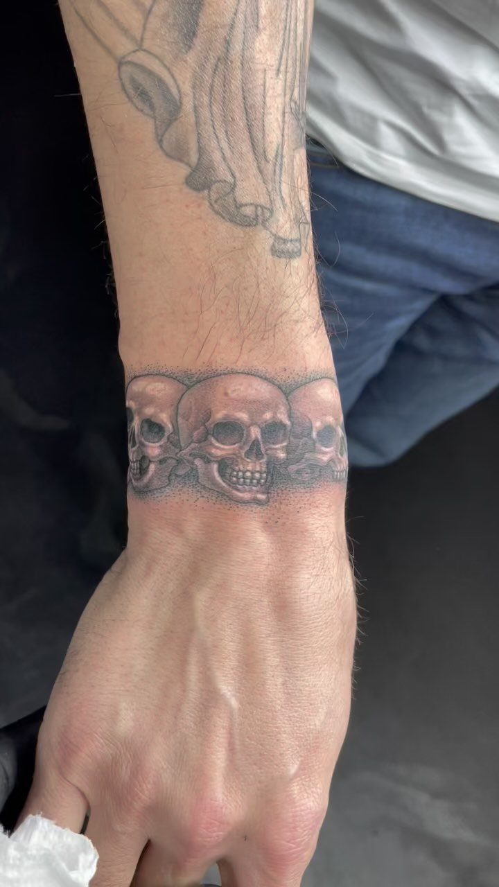 Man's wrist with a detailed black and grey tattoo of three human skulls.