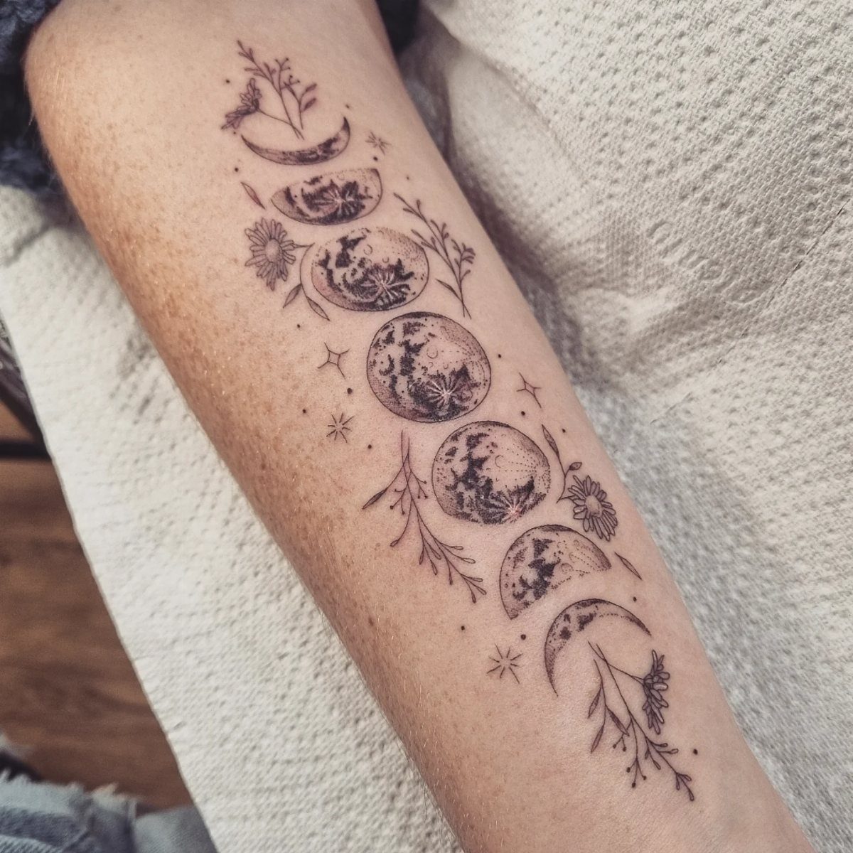 Moon phase tattoo on a forearm decorated with floral elements, stars, and a butterfly.