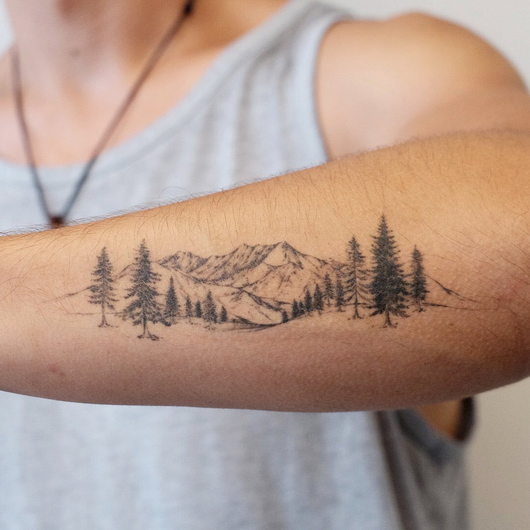 Mountain range tattoo with pine trees on a person's forearm in black ink