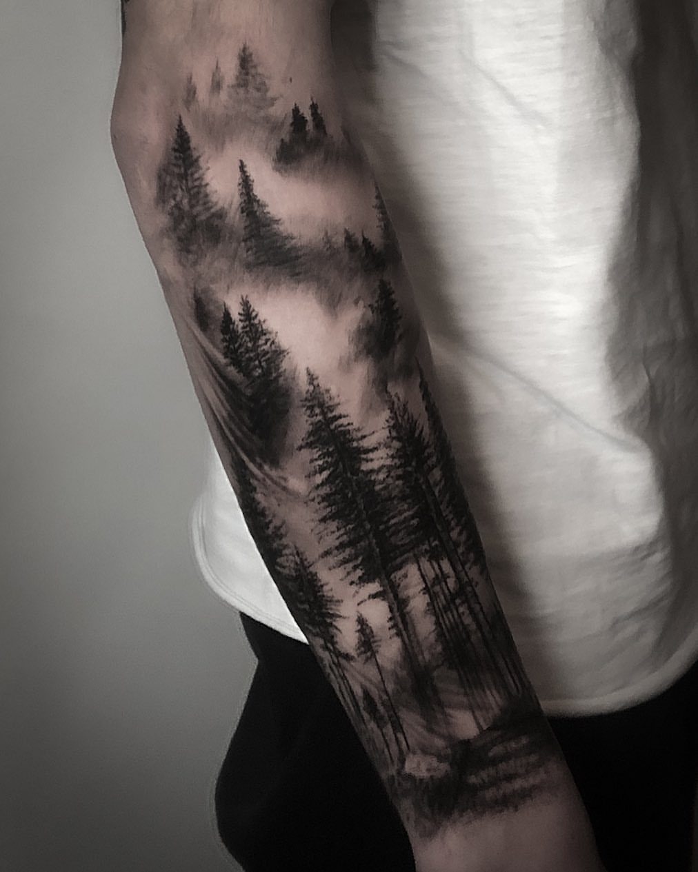 Forearm tattoo of a pine forest with misty negative space between dark trees.