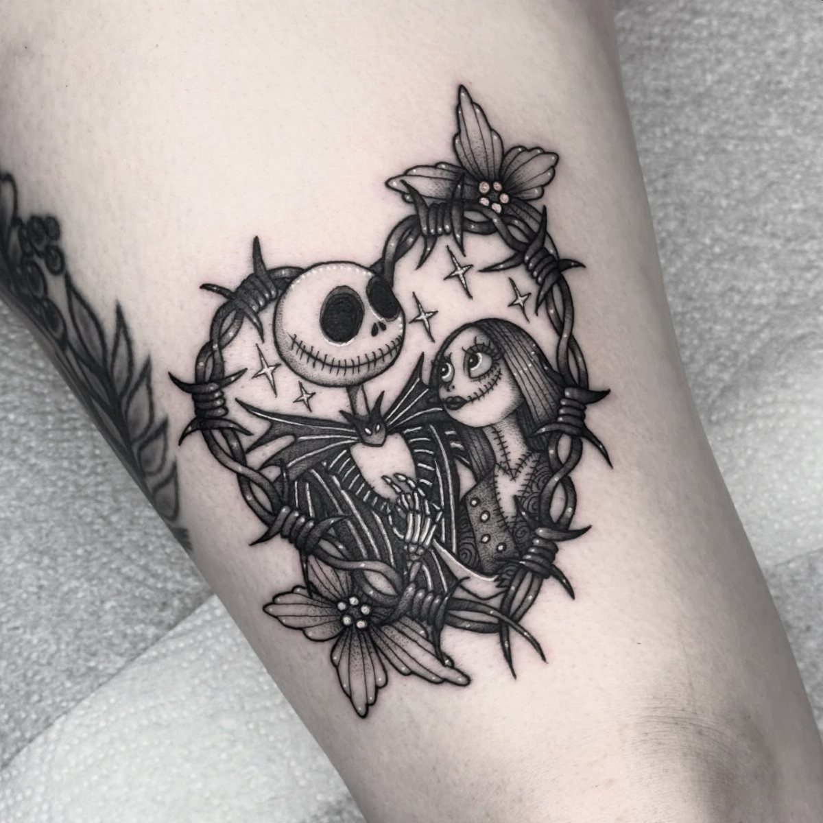 Jack and Sally tattoo enclosed in a barbed wire heart with small flowers.
