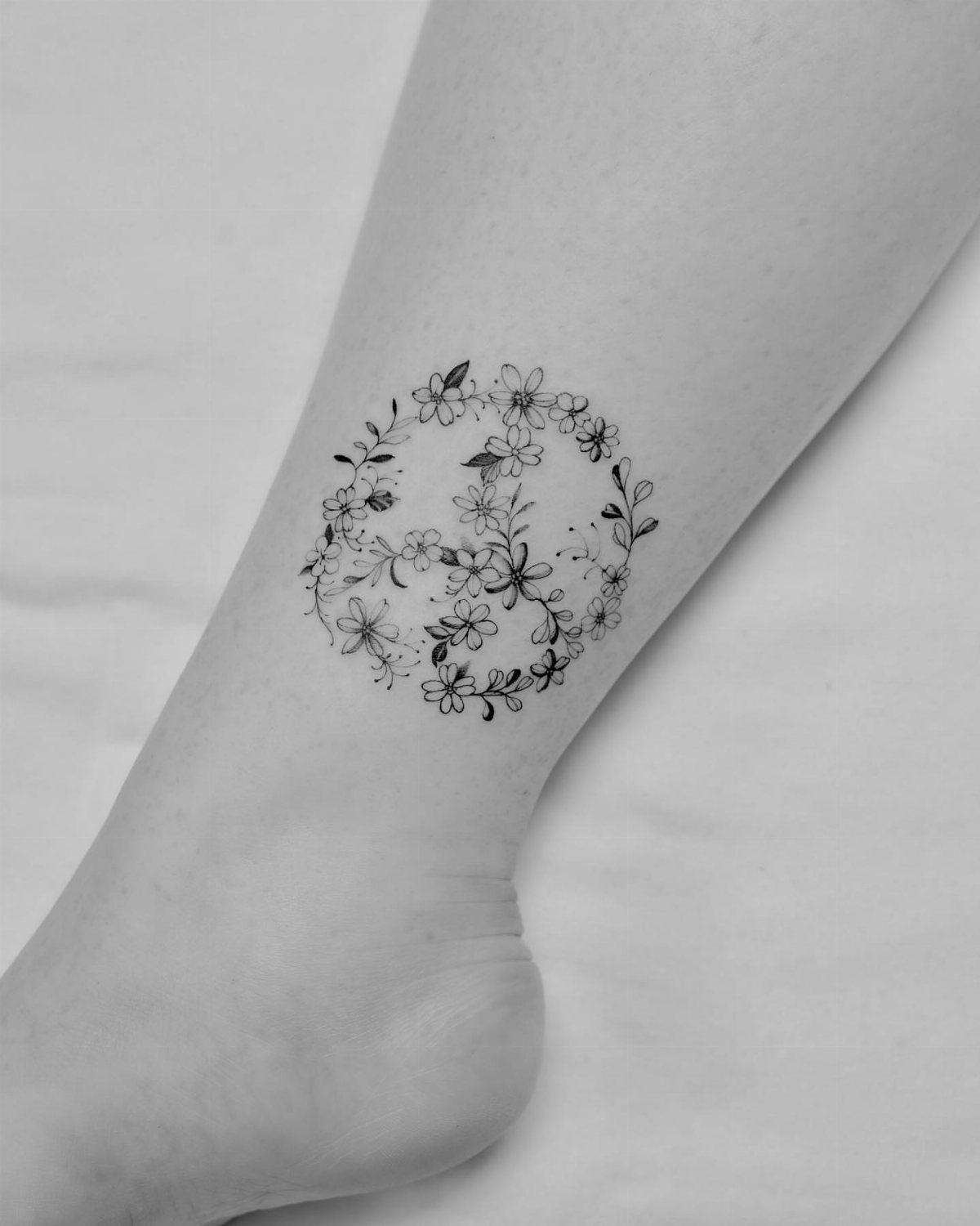 Peace symbol tattoo made of small flowers and vines on a person's ankle.