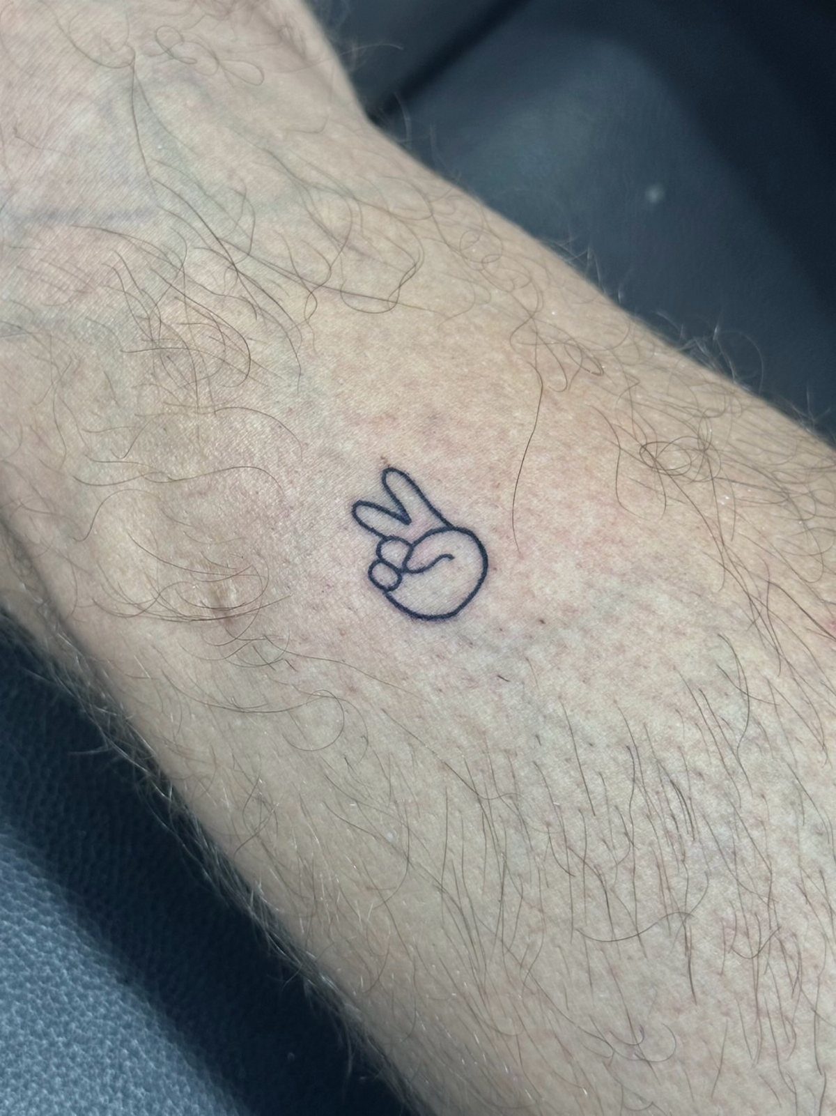 Hand peace sign tattoo outline on a person's hairy forearm.