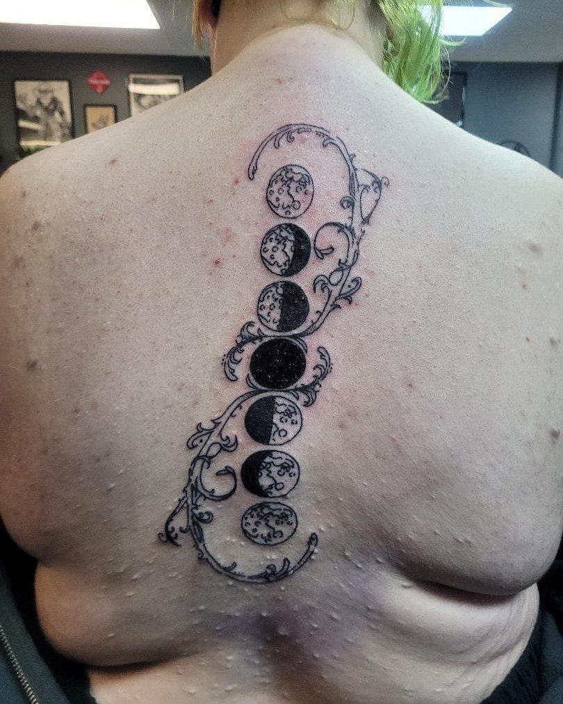 Phases of the moon tattoo with decorative swirls on a person's upper back.