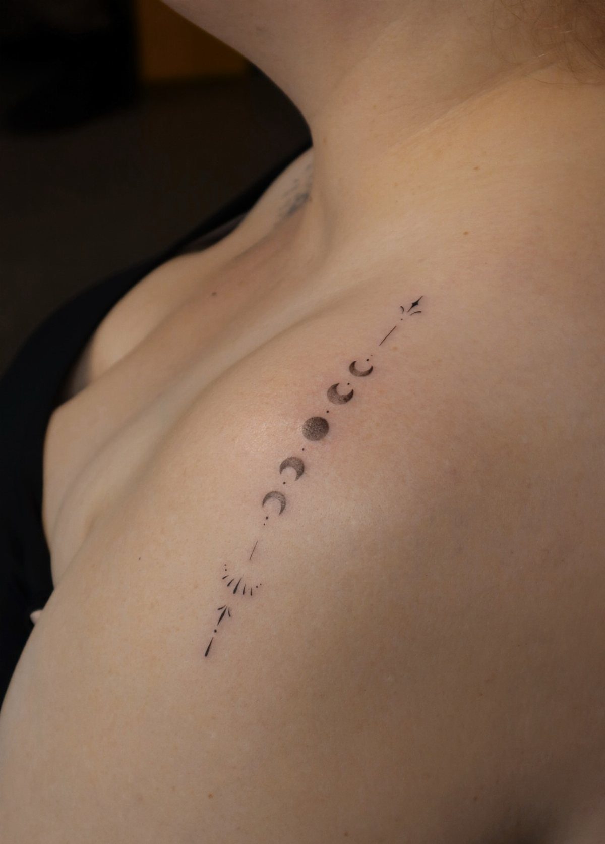 Minimalist moon phases tattoo in black ink on a person's shoulder and chest.