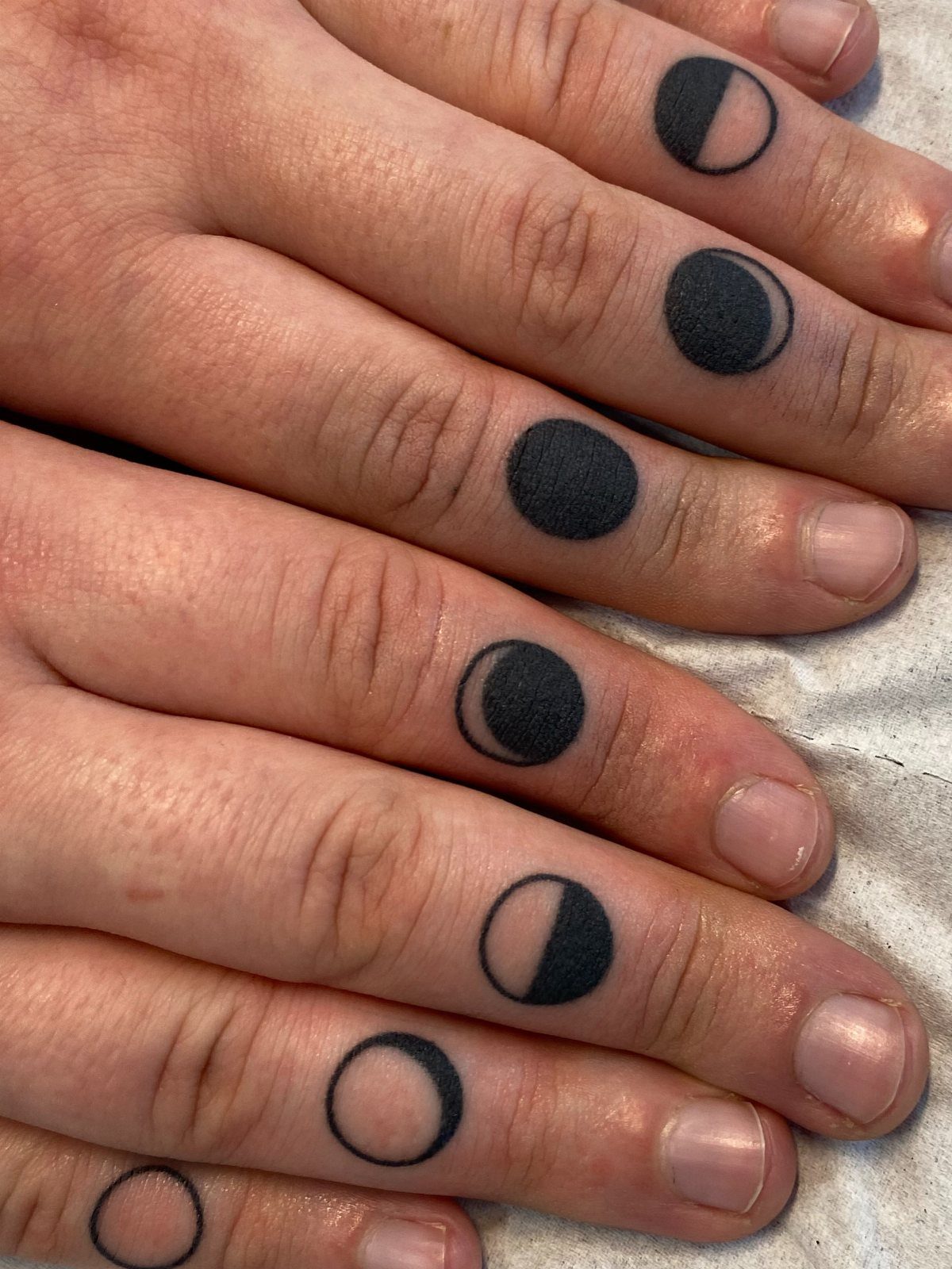 Moon phase tattoos in black ink on the fingers of two hands.