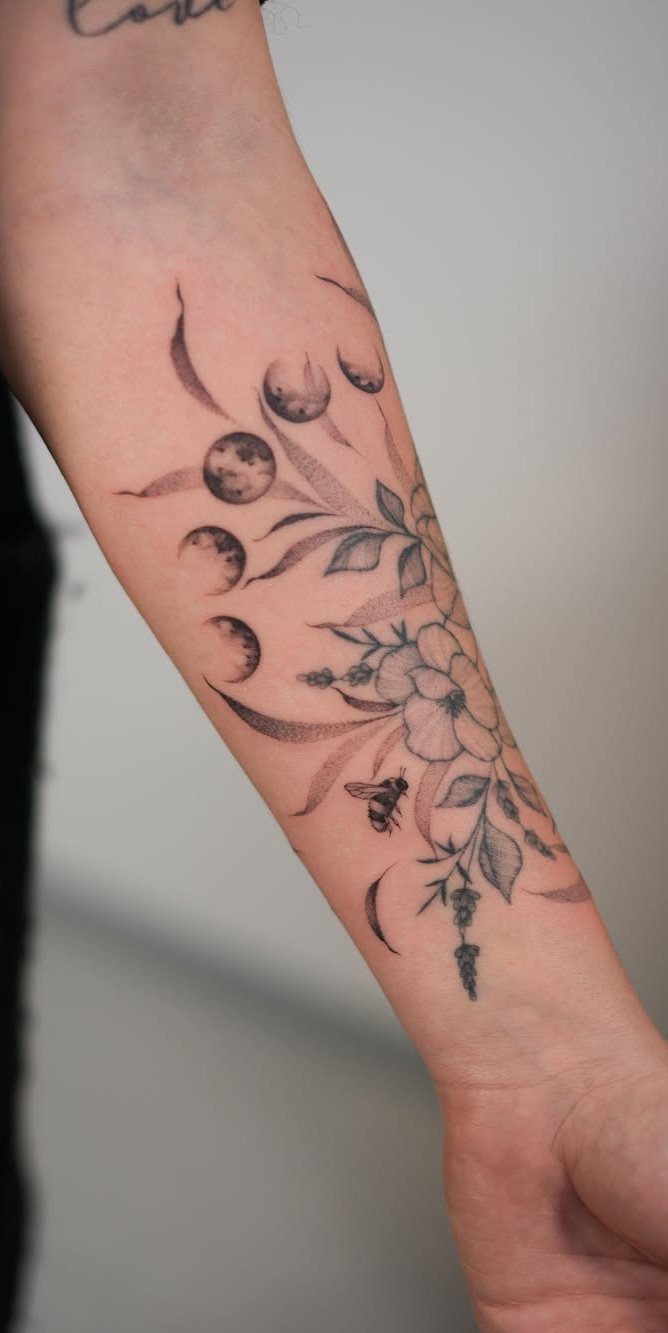 Forearm tattoo showing moon phases combined with delicate leaves, a flower, and a bee.