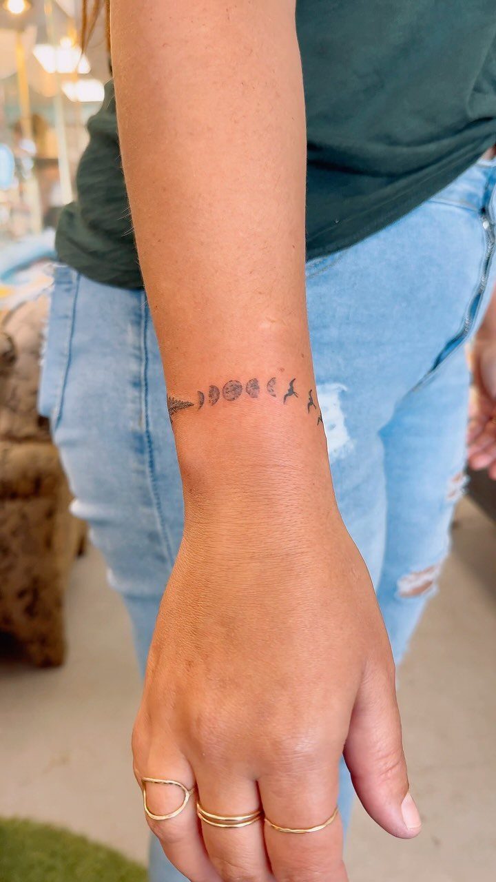 Moon phases tattoo wrapped around a person's wrist with gold rings on fingers.