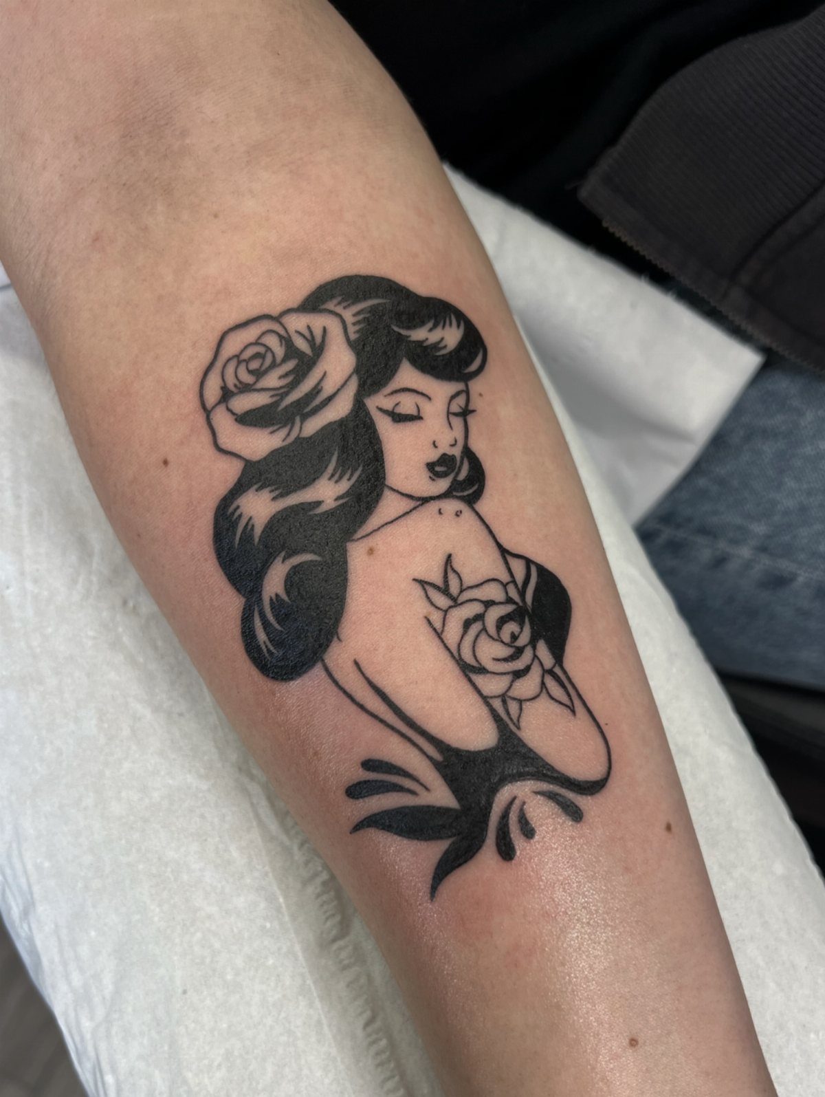 Pin up woman tattoo with roses on a person's forearm in black ink.