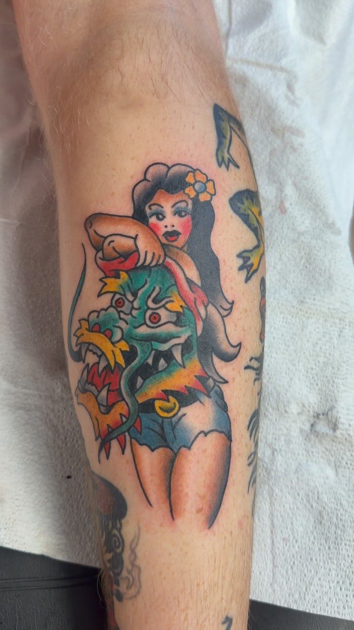 Pin-up woman tattoo leaning on a colorful dragon head on a person's leg