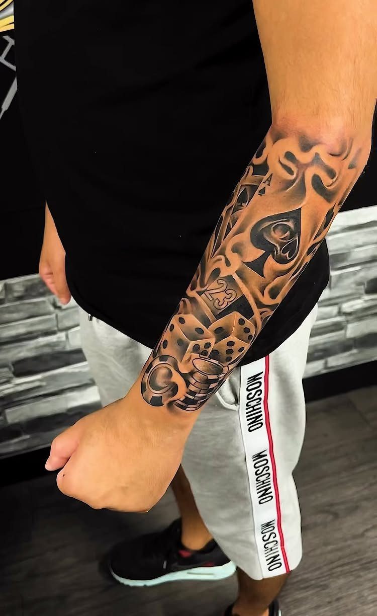 Forearm tattoo featuring an ace of spades, dice, and smoke in black and grey.