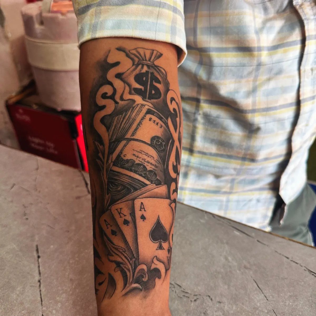Forearm tattoo featuring Ace of Spades playing cards, stacks of cash, and money bag.