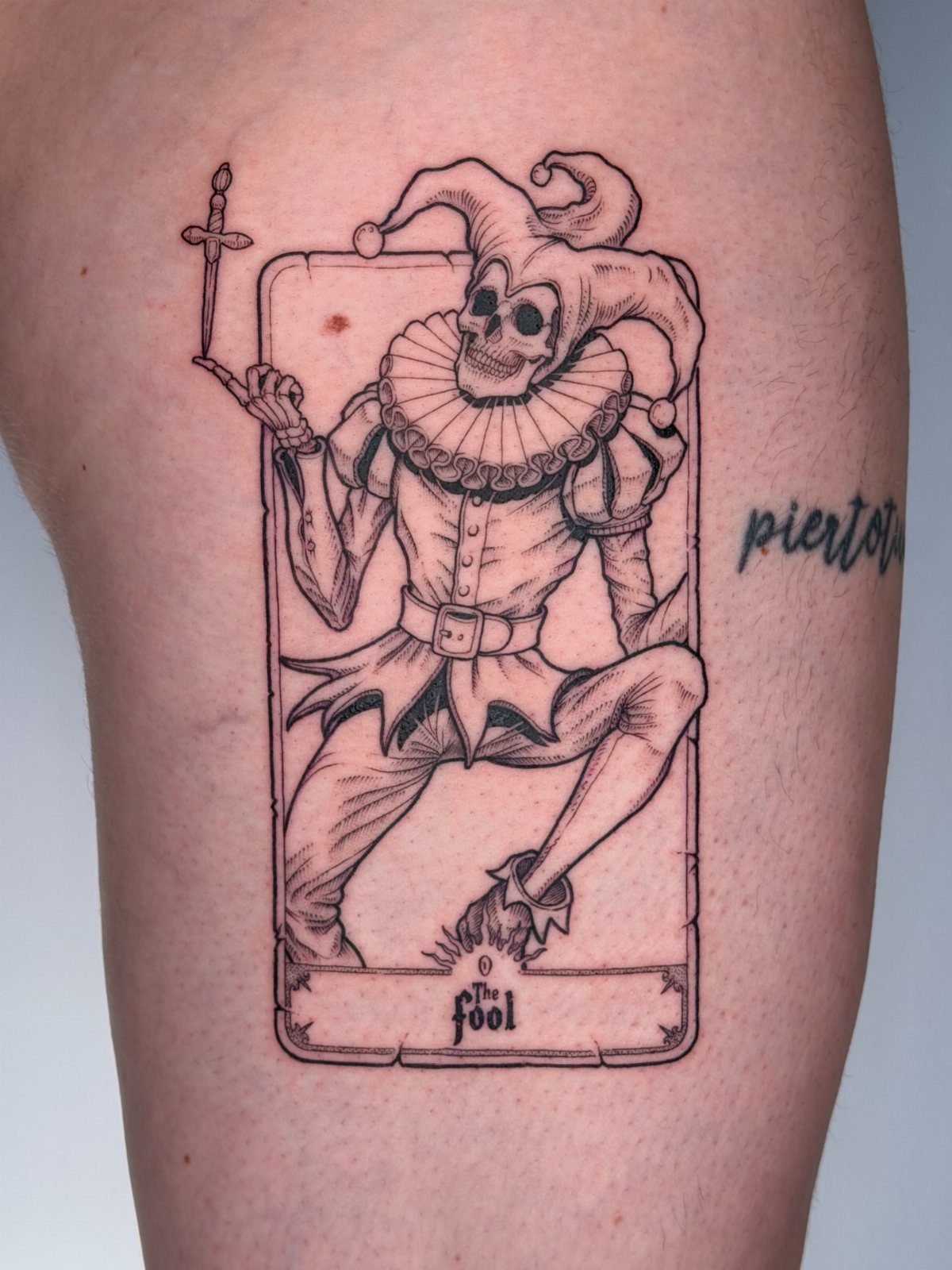 Skeletal jester playing card tattoo on a person's thigh with the text the fool