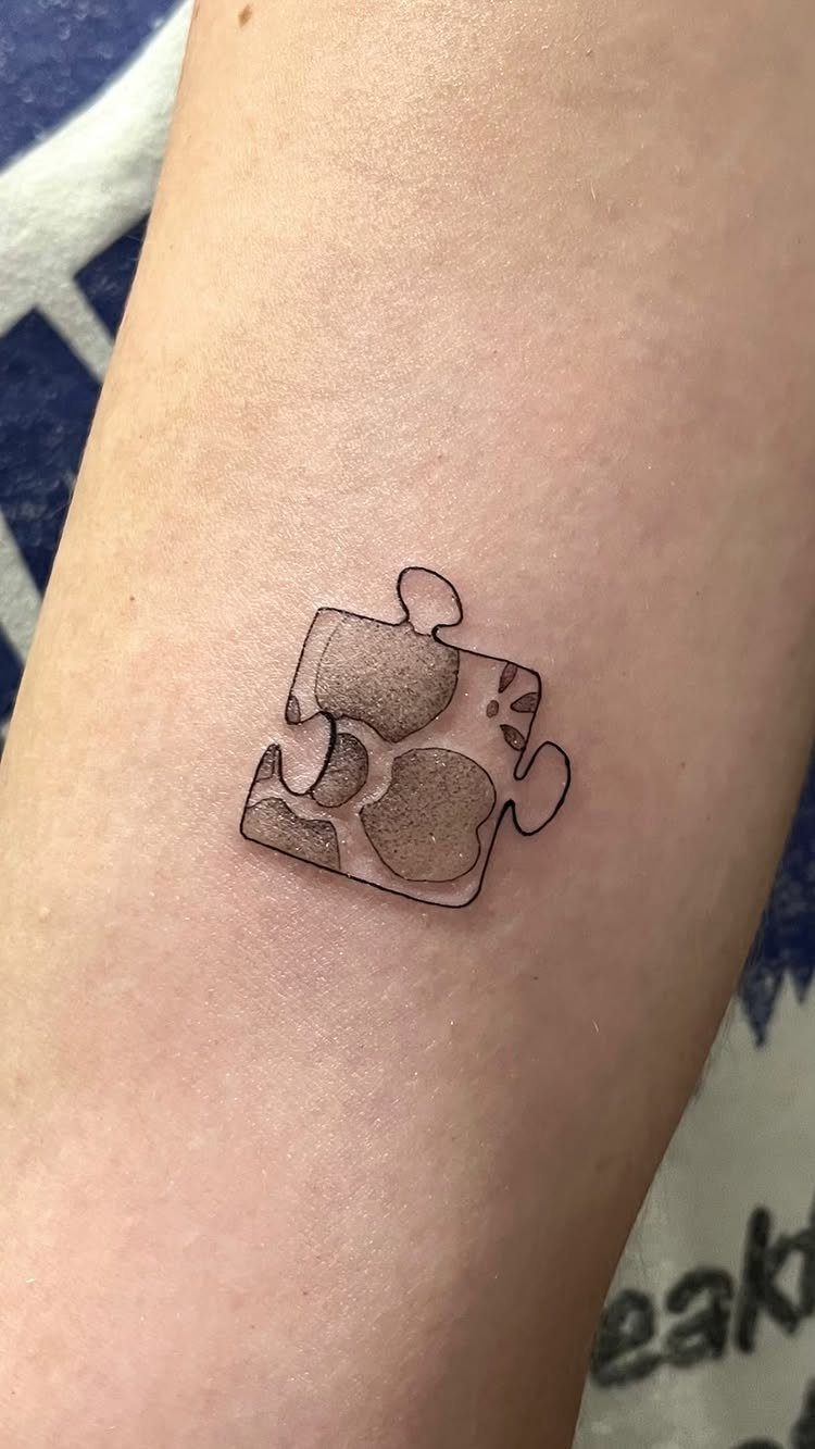 Single jigsaw puzzle piece tattoo with gray shading on a person's forearm.