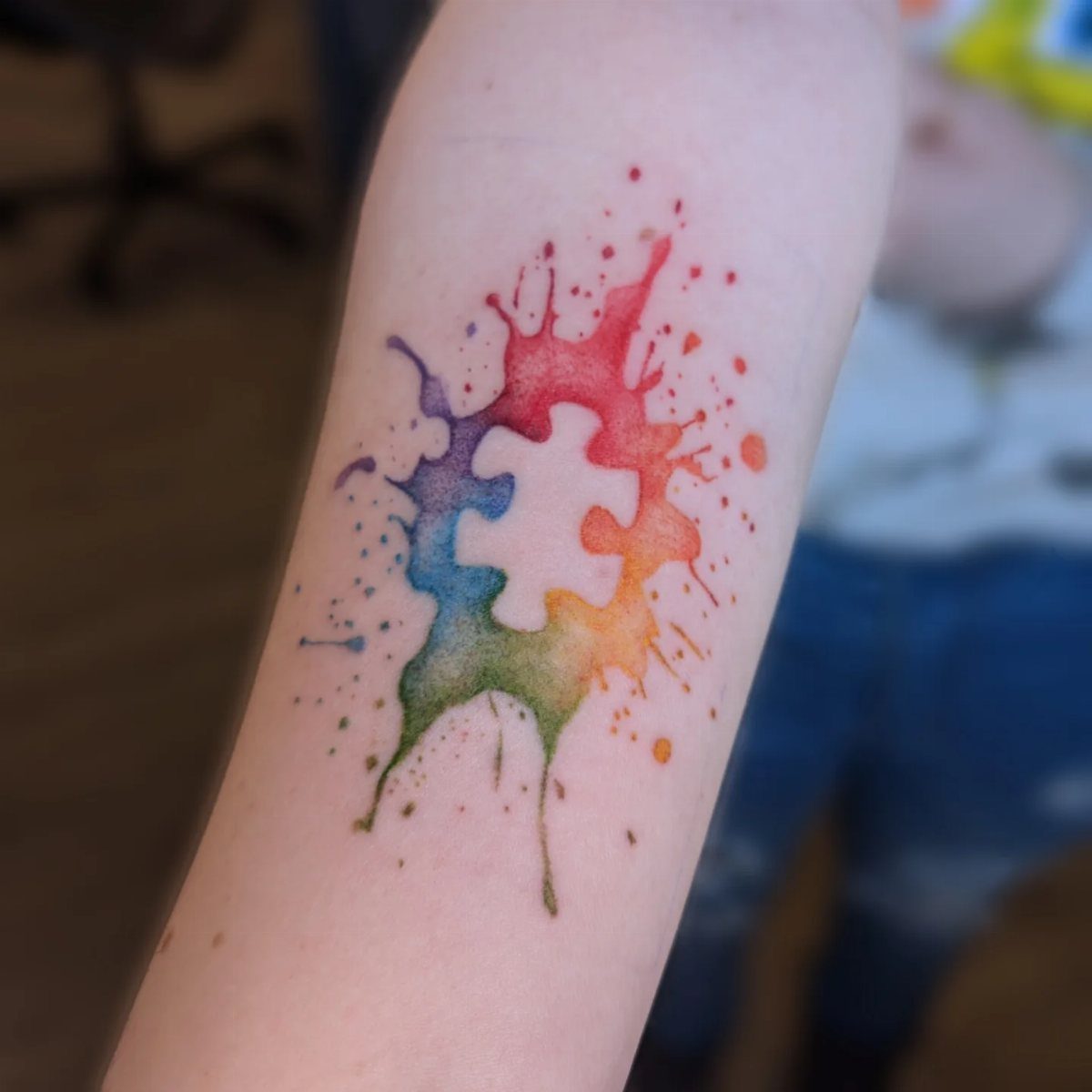 Watercolor tattoo on a forearm showing a rainbow paint splash forming a puzzle piece