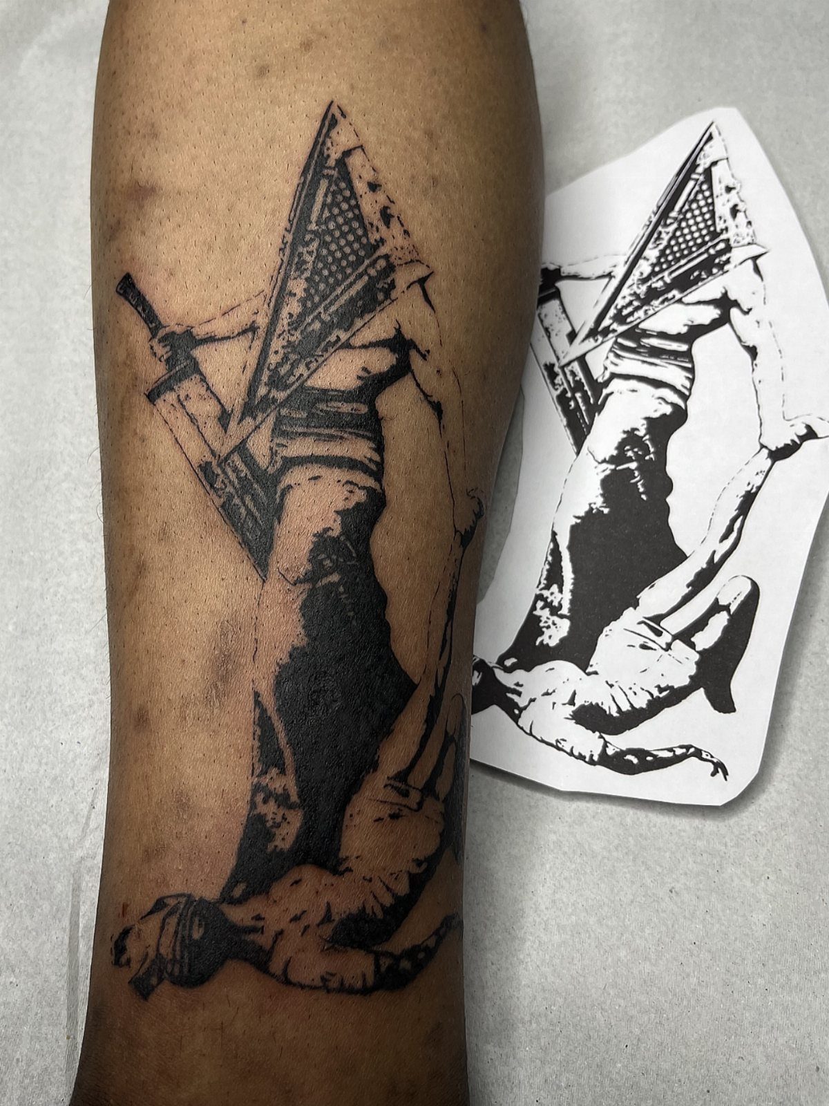 Pyramid Head tattoo on an arm with the original stencil design beside it.