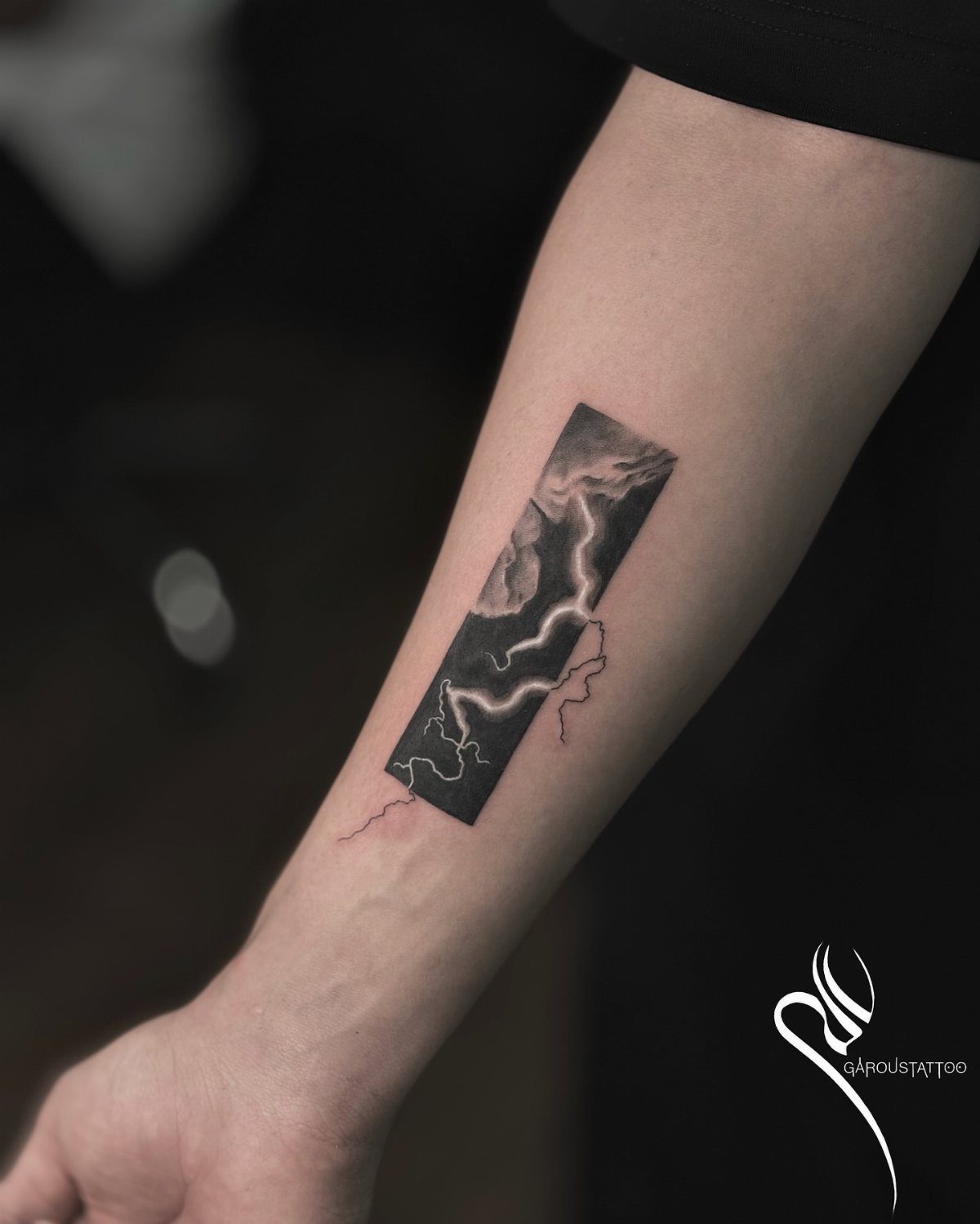 Forearm tattoo of a rectangular frame showing dark clouds and white lightning bolts.