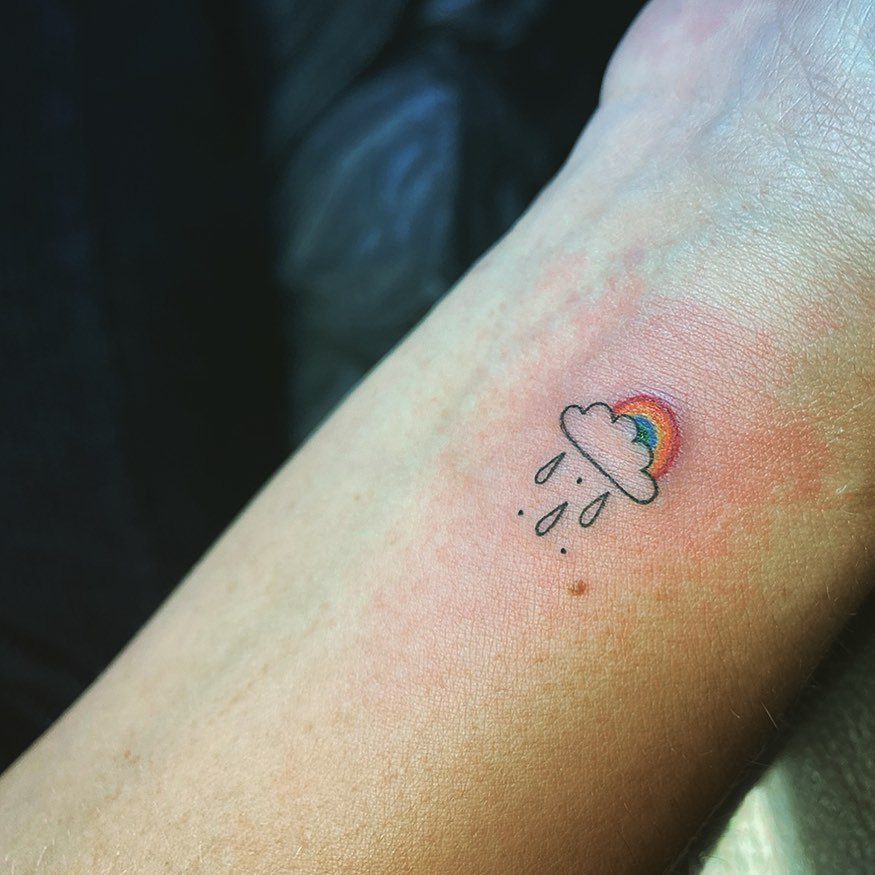 Cloud tattoo with rain droplets and a colorful rainbow on a forearm.