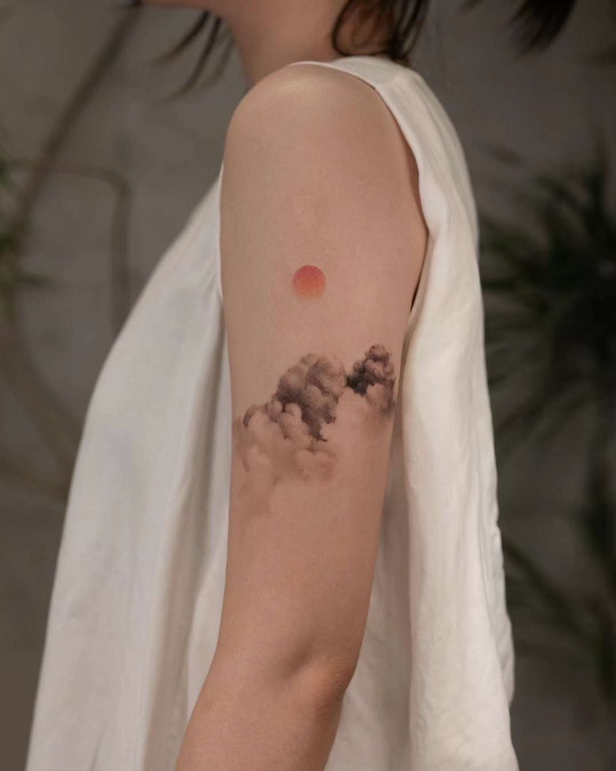 Tattoo of shaded clouds and a small red sun on an upper arm.