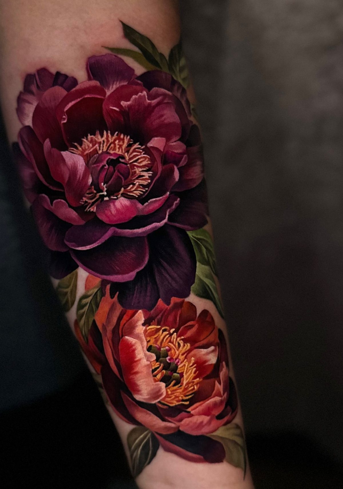 Realistic peony tattoo in purple and red with green leaves on an arm.