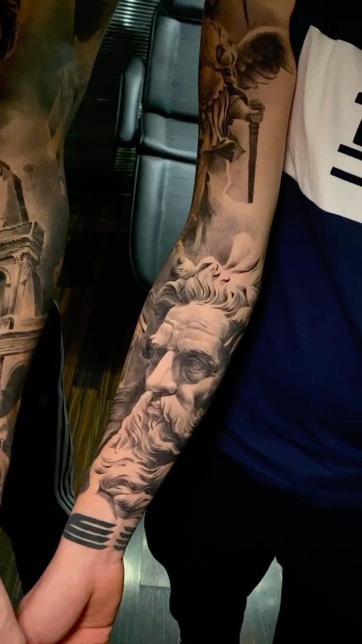 Realistic black and grey tattoo of a classical statue face on a forearm.