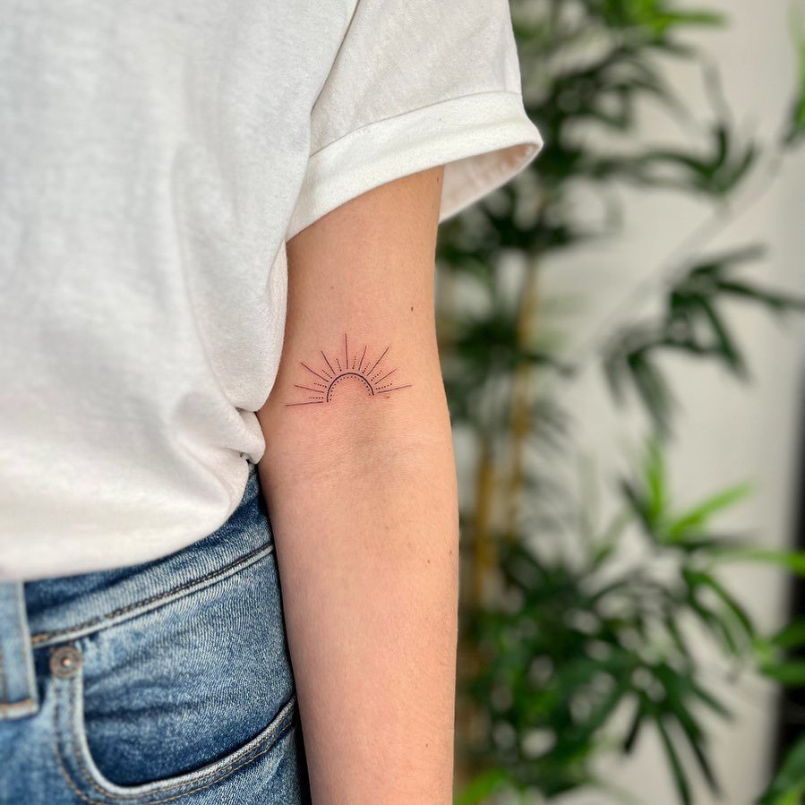 Minimal rising sun tattoo with thin lines on a person's upper arm.