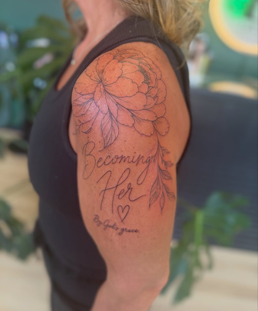 Tattoo on upper arm with a peony flower and text Becoming Her.