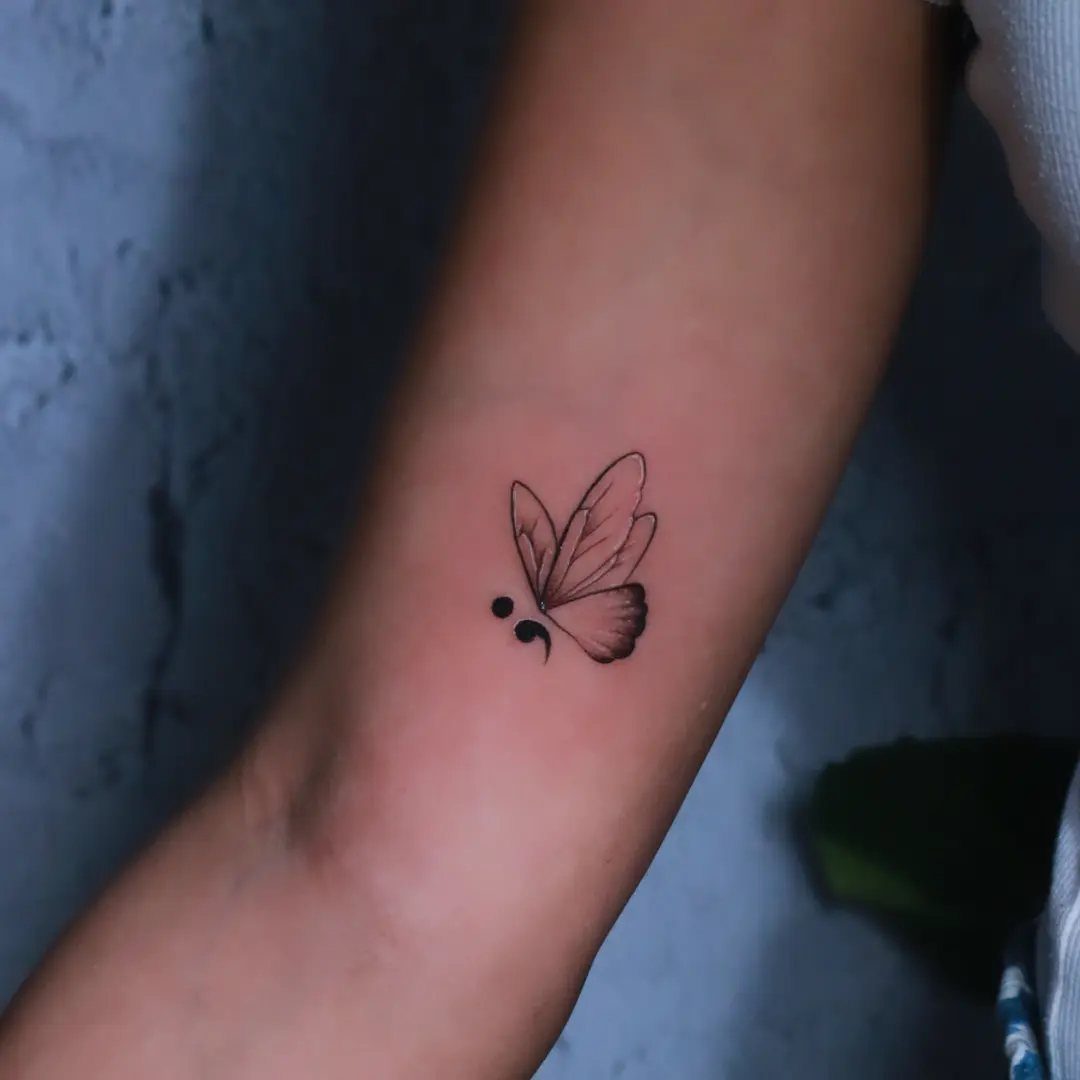 Butterfly tattoo with a semicolon integrated into the wing on a forearm